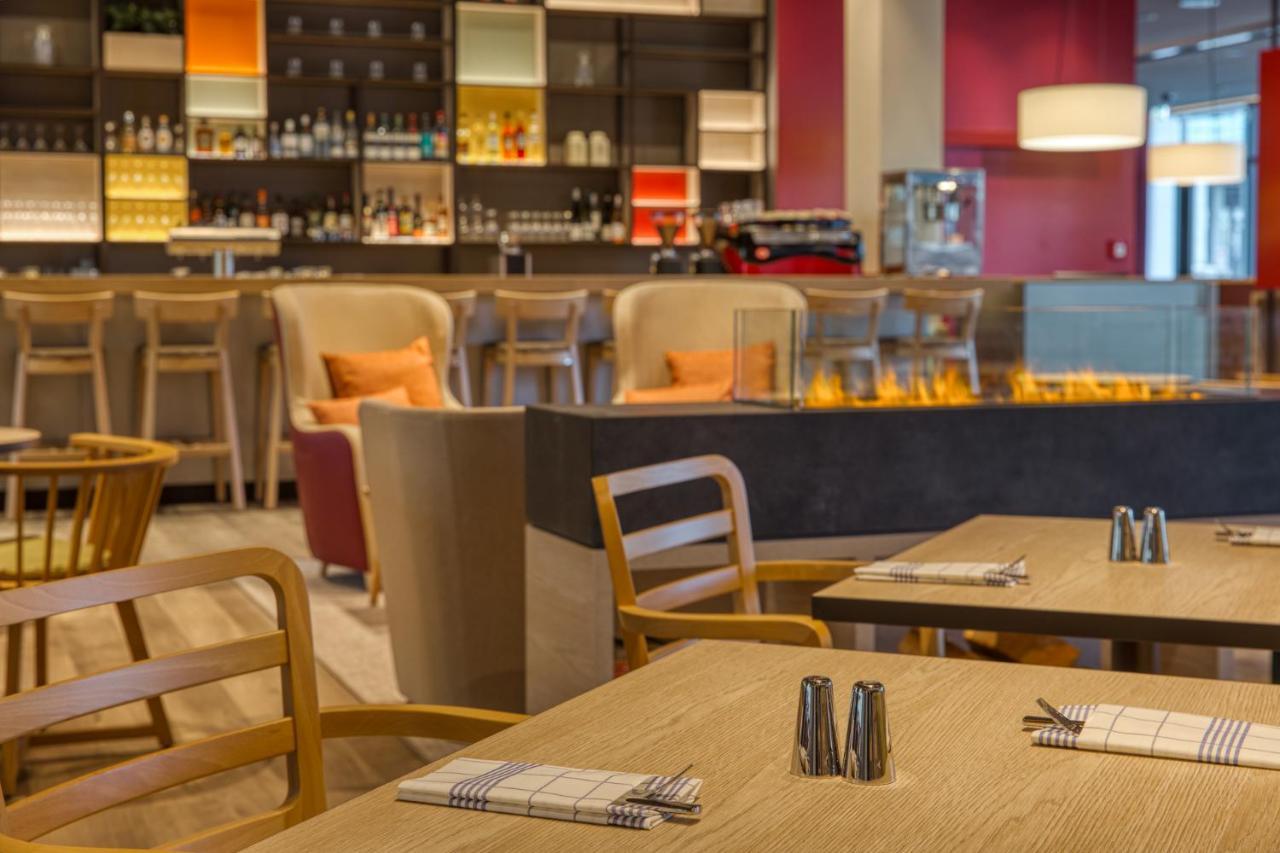 Intercityhotel Berlin Airport Ber Terminal 1 & 2 Schoenefeld Exterior photo The photo shows a modern restaurant or bar interior. In the foreground, there are wooden tables set with napkins and condiments. The seating includes wooden chairs and plush seating in various colors in the background. A stylish bar area is visible w